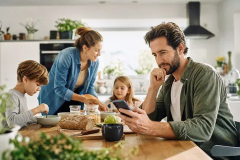 Symbolbild zum Ratgeber „Dynamischer Stromtarif: Rechnet sich das für dich?“: eine Familie beim Frühstück in der Küche, ein Elternteil prüft nachdenklich das Smartphone.